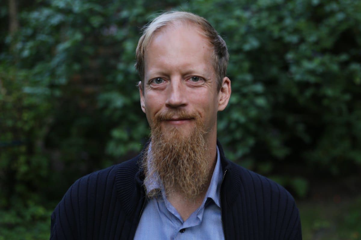 Juuso Kangas hymyilee, taustalla vihreää lehvästöä.